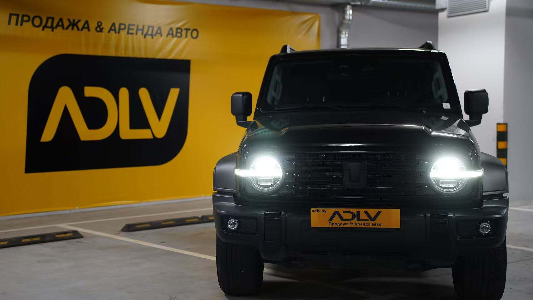 TANK 300 с включенными фарами на фоне баннера с логотипом ADLV в паркинге.