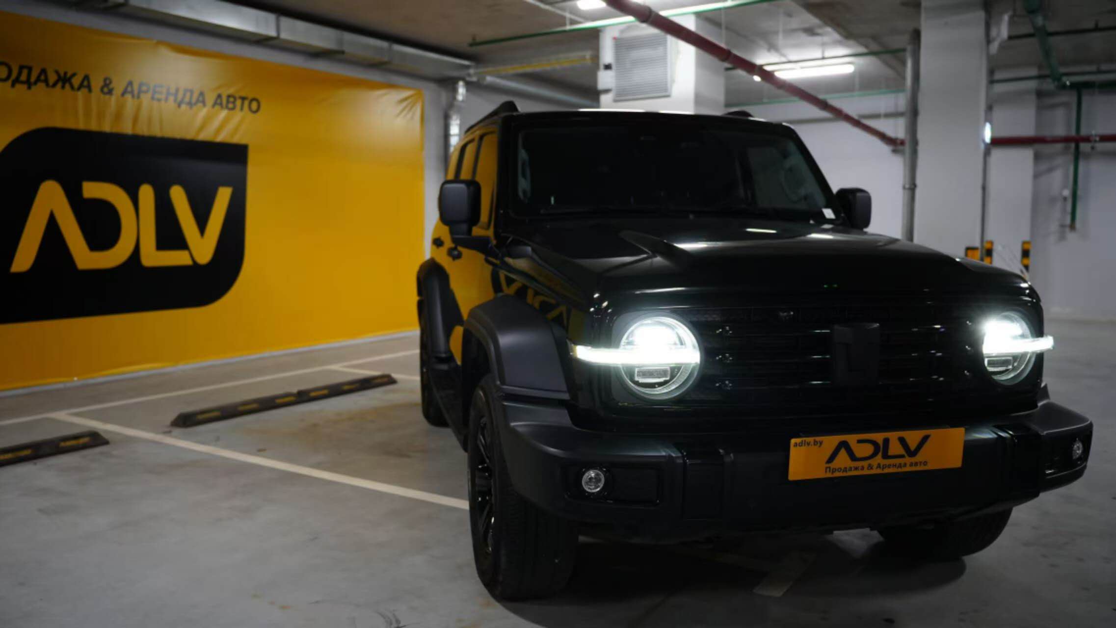TANK 300 с включенными фарами на фоне баннера с логотипом ADLV в подземном паркинге.